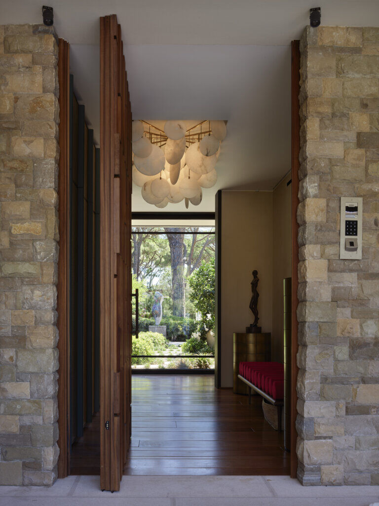 Entrance Framed by Stone and Light — Saint-Tropez Villa Entrance framed by stone walls and wooden panels, opening toward a garden with a sculpture in Primadesign’s Saint-Tropez villa.